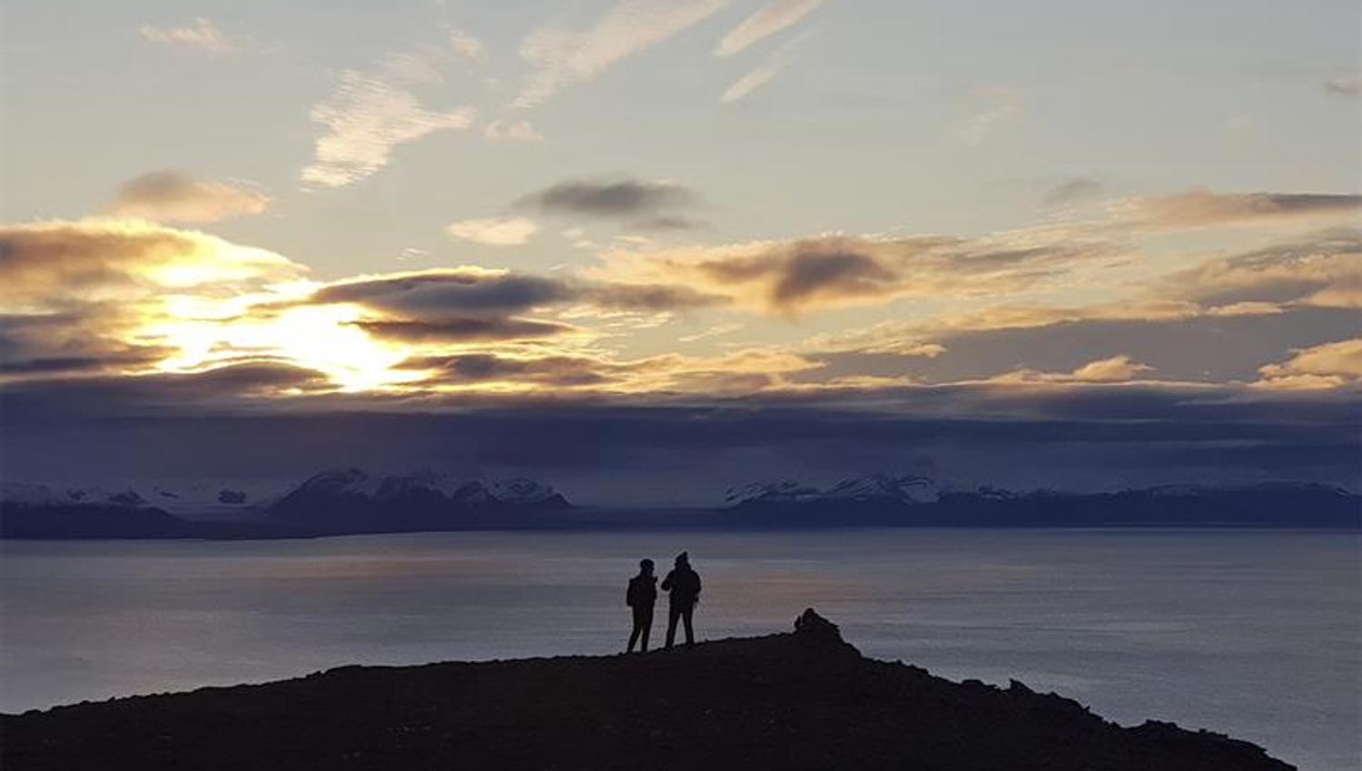 To personer på en fjelltopp i silhuett mot en mørk fjord og en solnedgang bak lave skyer i bakgrunnen
