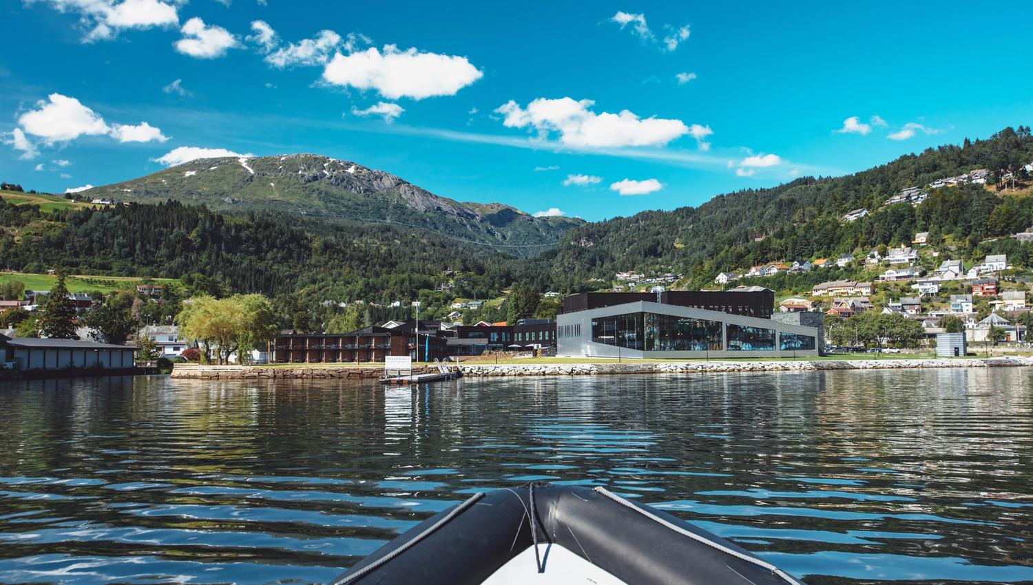 View of Hardangerfjord Hotel from the fjord, surrounded by mountains and a charming village.