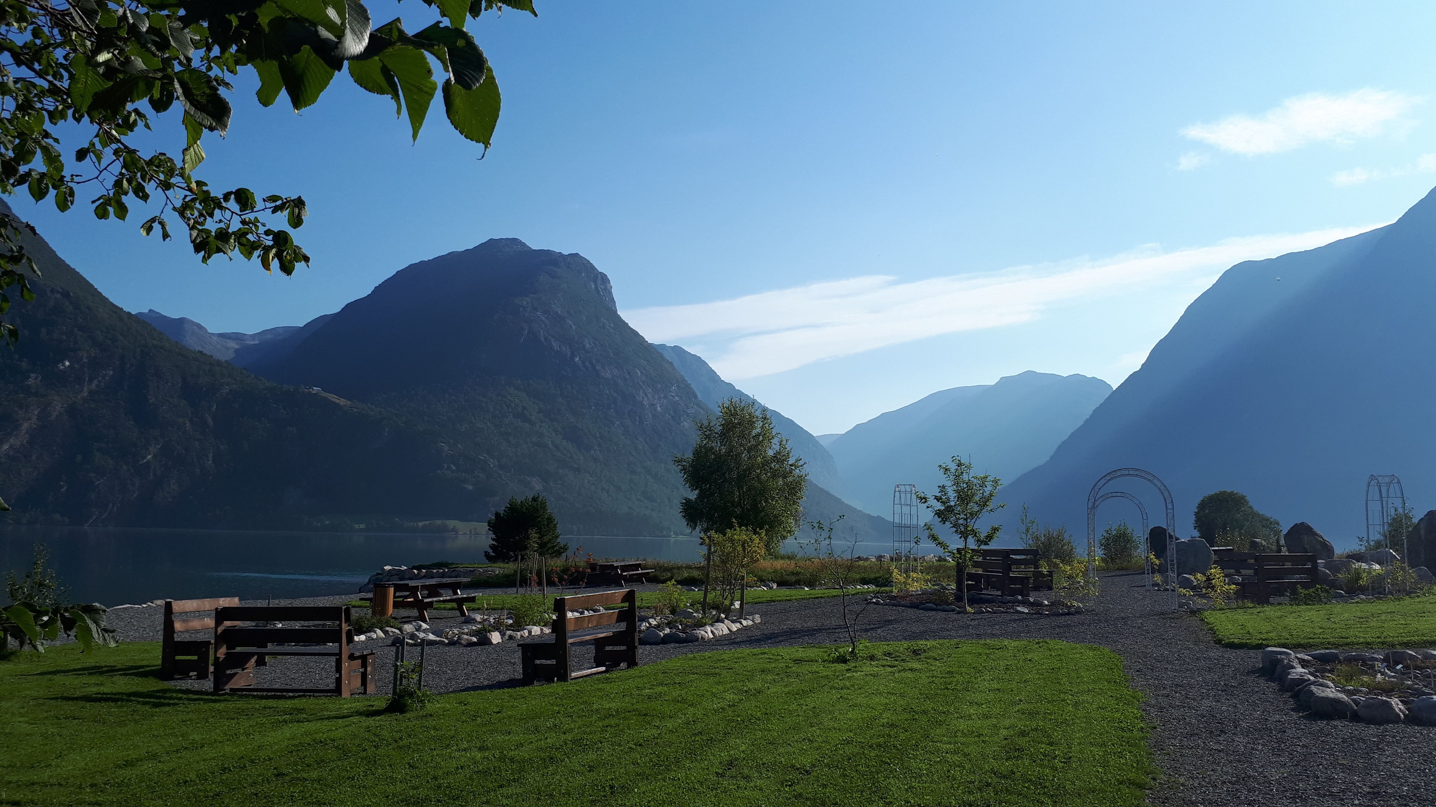 Grøn park med grusgangar og benker ved ein stille fjord, med store fjell og blå himmel i bakgrunnen..