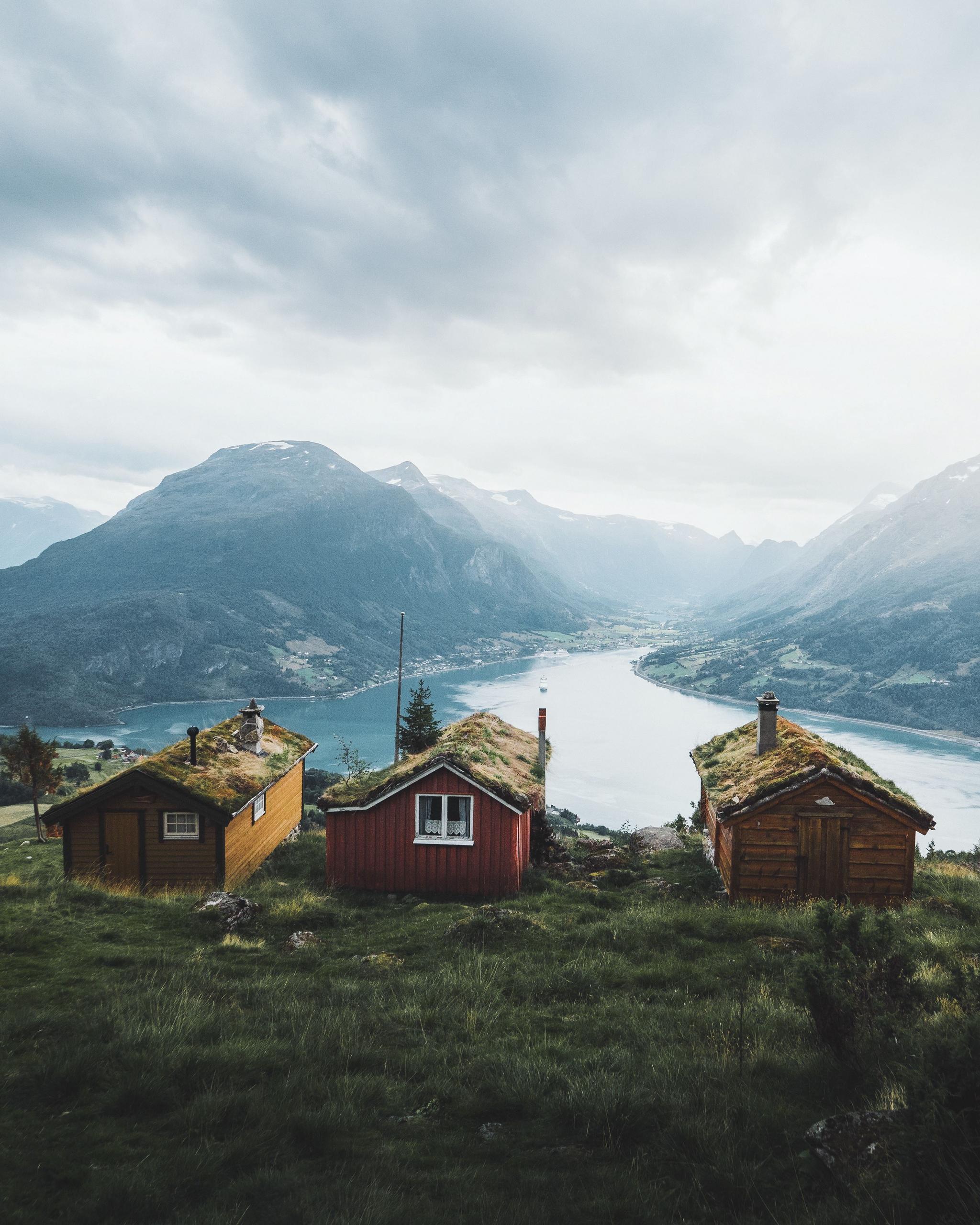 Three small wooden cabins with grass roofs sit on a hillside above a fjord, with mountains and cloudy sky beyond.