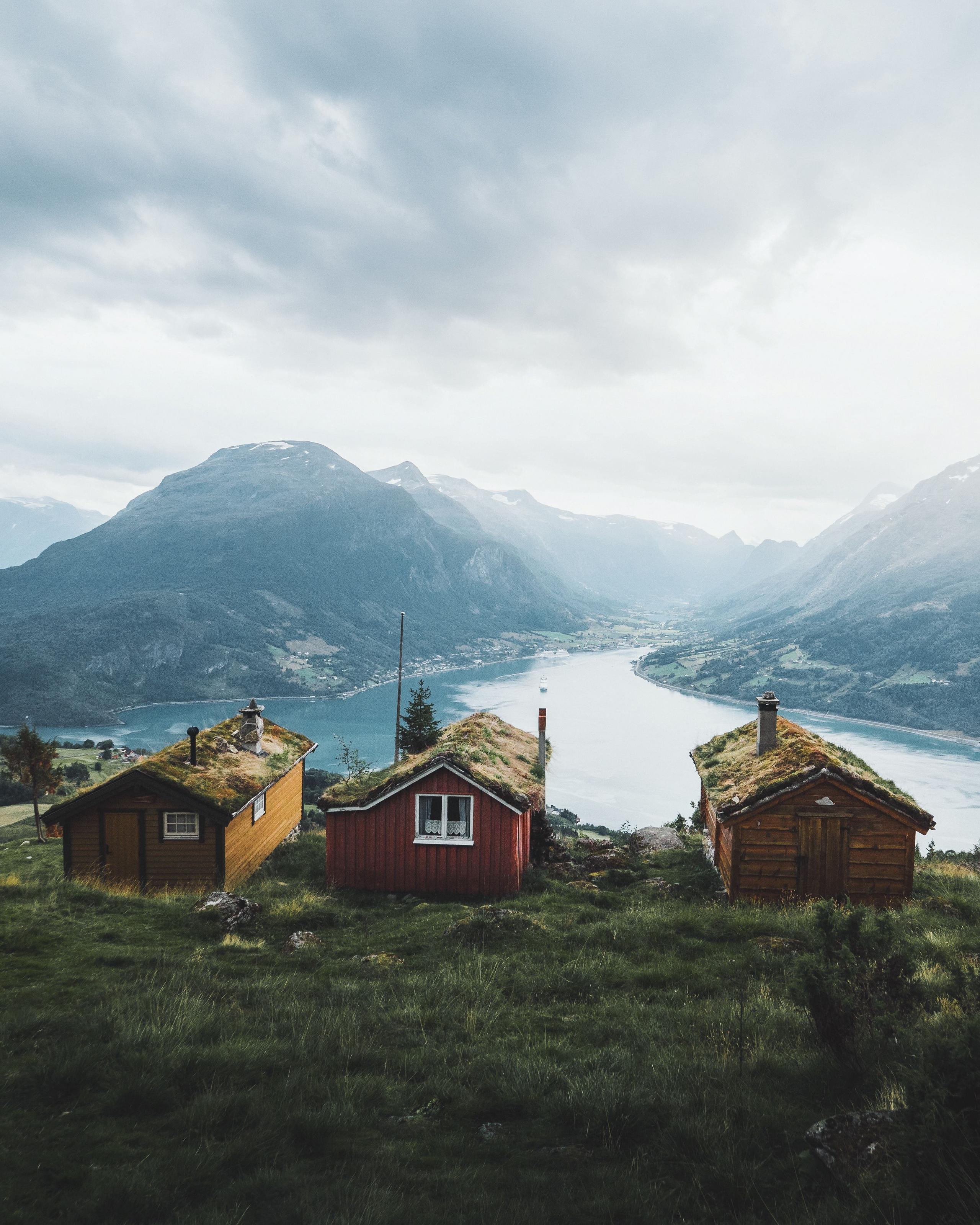 Three small wooden cabins with grass roofs sit on a hillside above a fjord, with mountains and cloudy sky beyond.