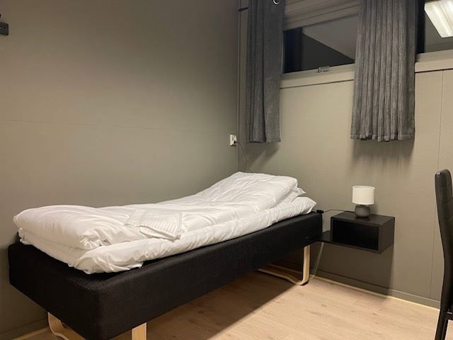 A single bed with white bedding sits in a gray room beside a floating nightstand and a curtained window.