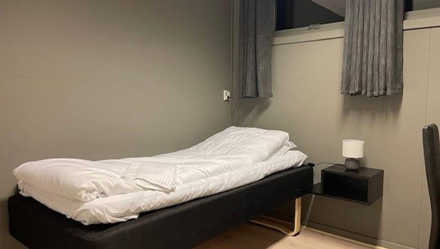 A single bed with white bedding sits in a gray room beside a floating nightstand and a curtained window.