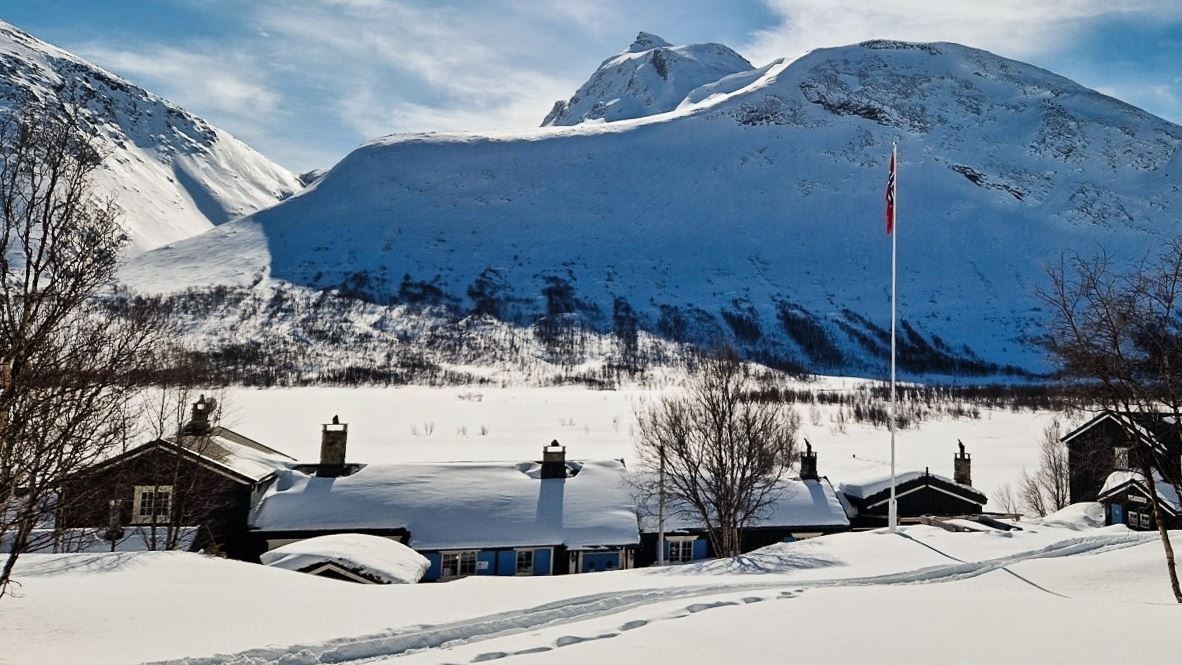 Snow-covered cabins and bare trees sit in a valley below steep white mountains under a partly cloudy sky..