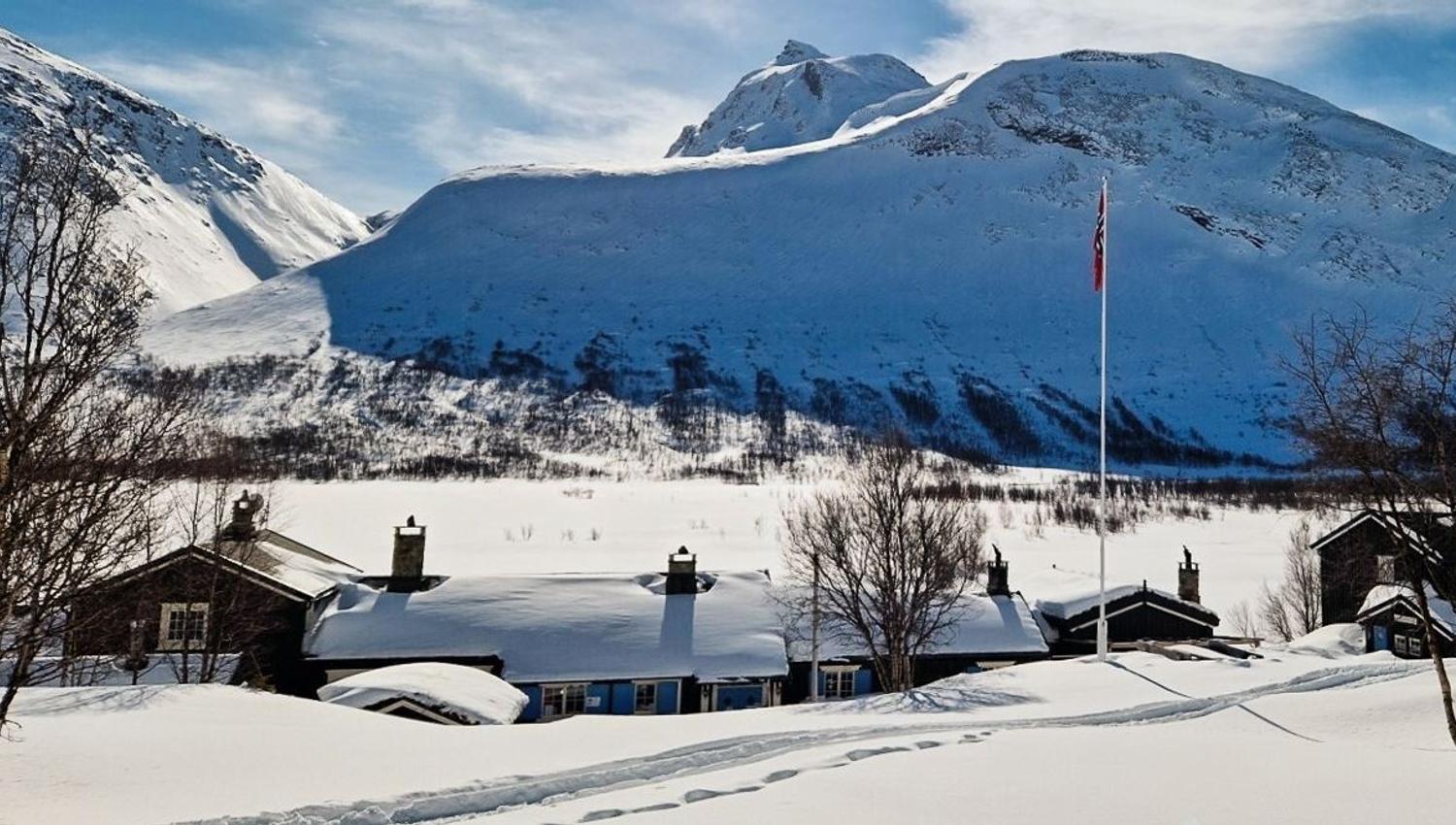 Snow-covered cabins and bare trees sit in a valley below steep white mountains under a partly cloudy sky..