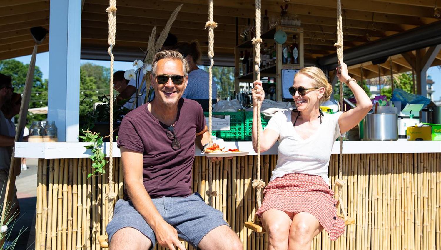 To personer sitter i husker ved en strandbar under et stråtak.