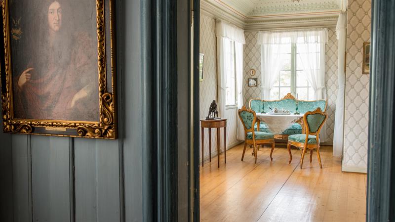 Interior of a historic home with light-coloured walls, antique furniture and a small table positioned by the window.