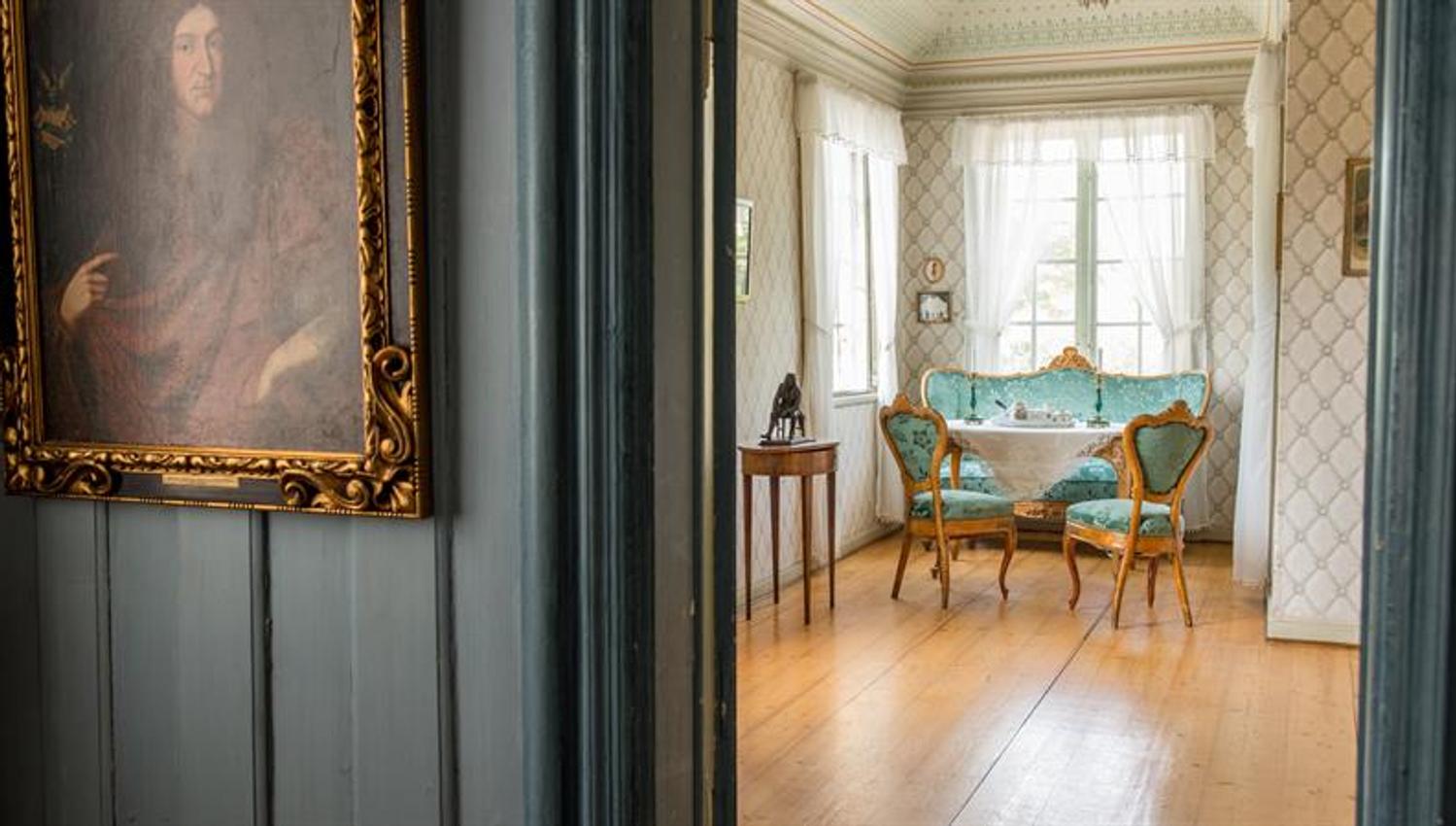 Interior of a historic home with light-coloured walls, antique furniture and a small table positioned by the window.