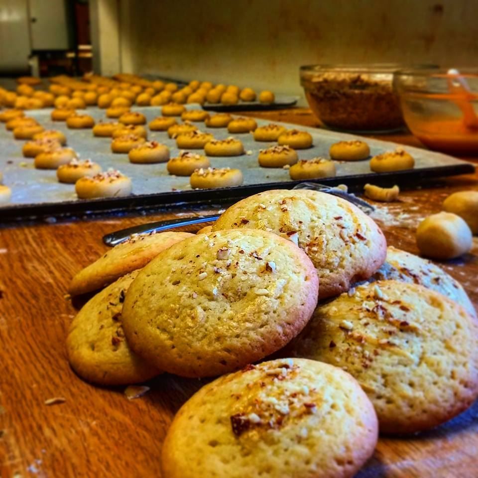 Close-up of baked cookies on a wooden counter, with trays of unbaked dough balls and mixing bowls in the background.