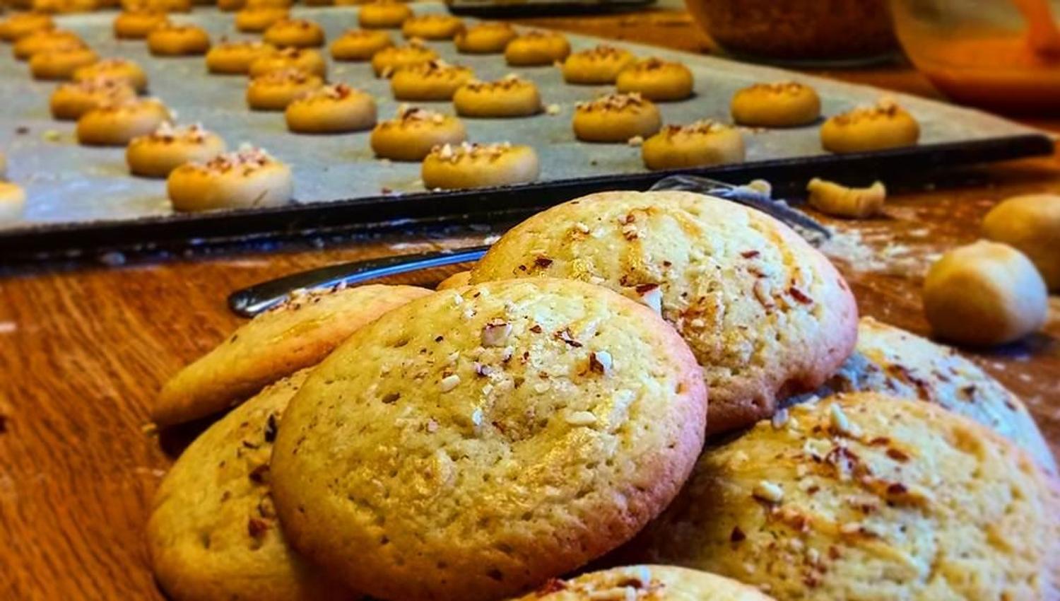 Close-up of baked cookies on a wooden counter, with trays of unbaked dough balls and mixing bowls in the background.