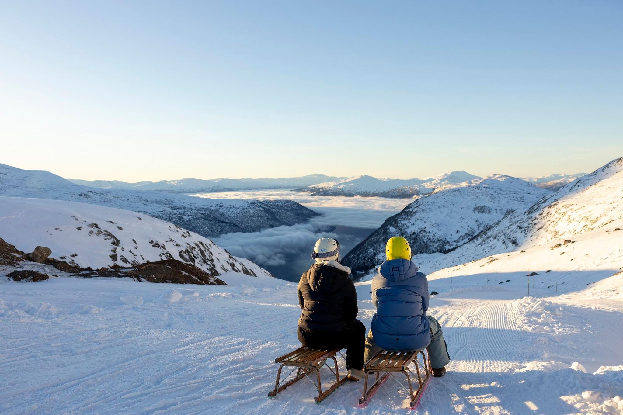 Two people in helmets sit on wooden sleds on a snowy slope, looking over mountains and a cloud-filled valley.