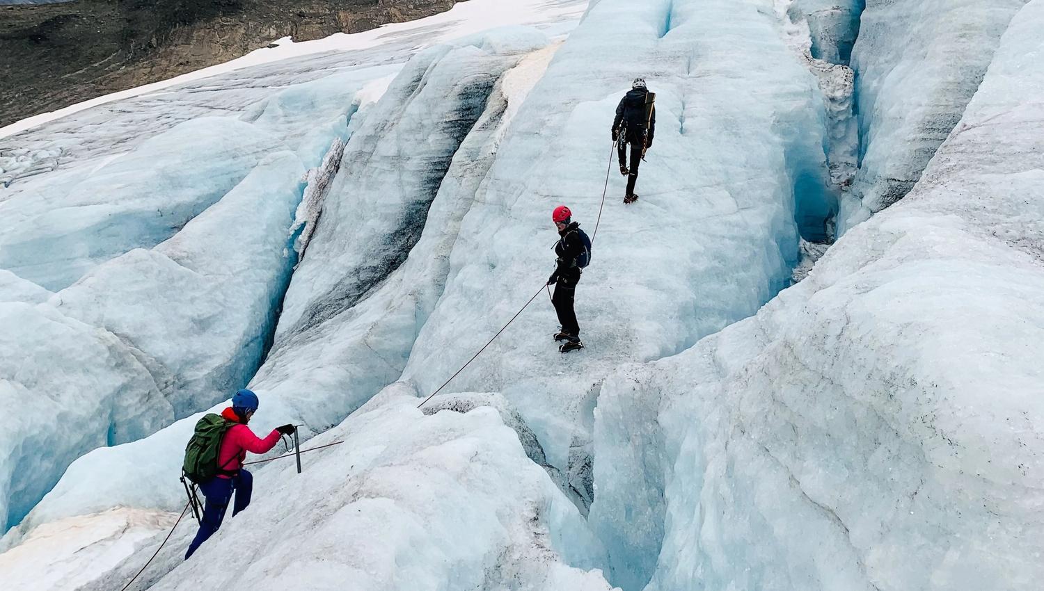 Three mountaineers roped together traverse a blue glacier with deep crevasses, wearing helmets and carrying packs..