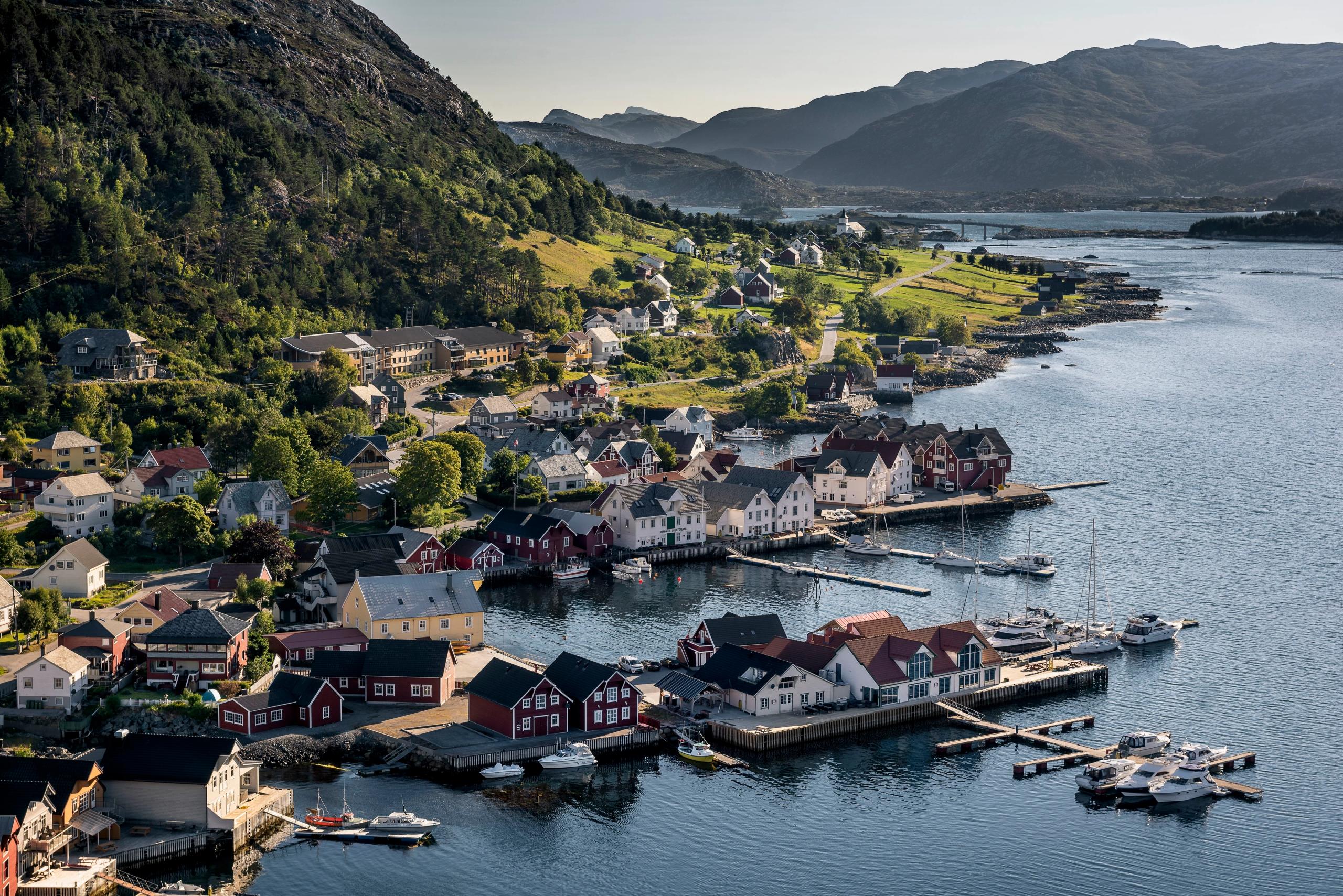Aerial view of a waterfront village with colorful houses, docks and boats beside calm water and green mountains.