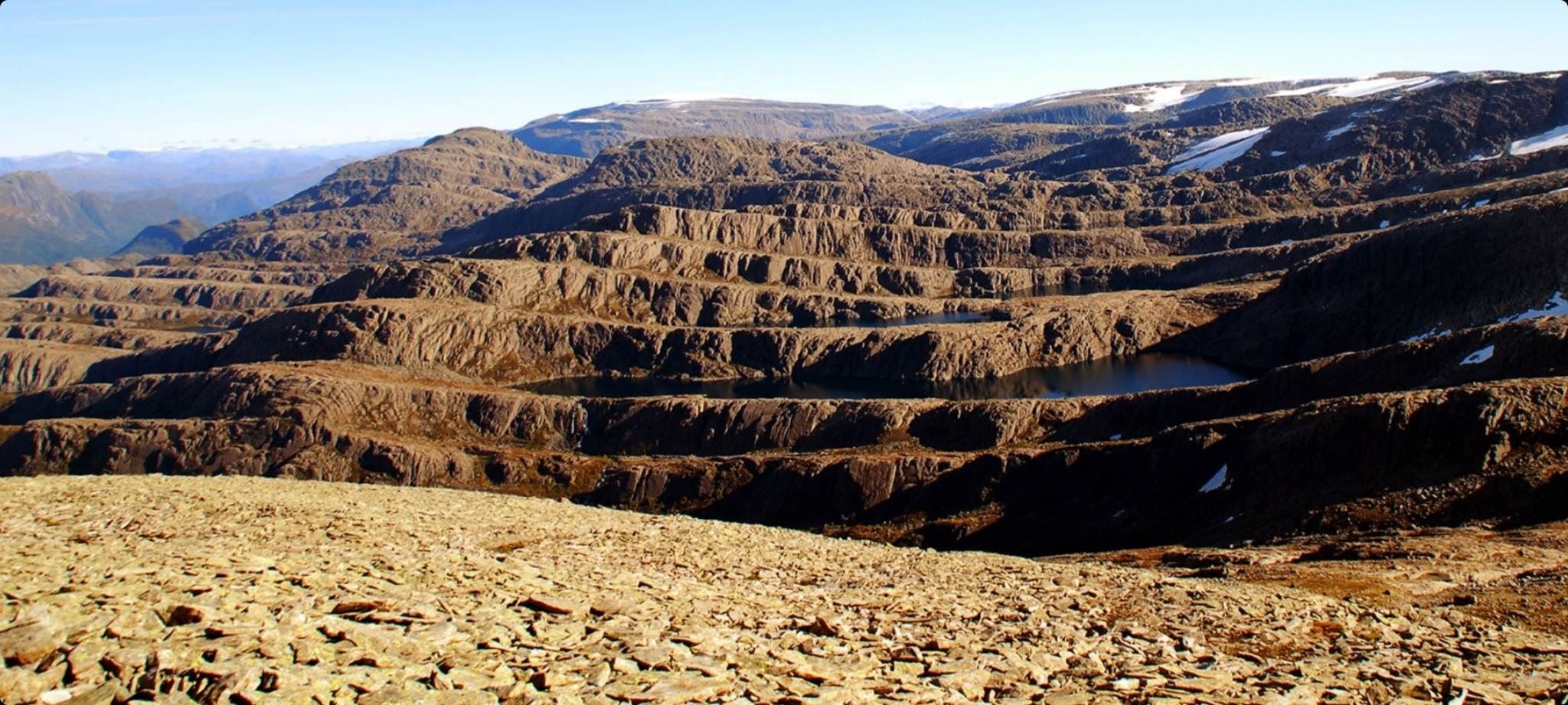 Panoramautsikt over bratt, steinete fjellandskap med små innsjøar og snøflekker under klar blå himmel..