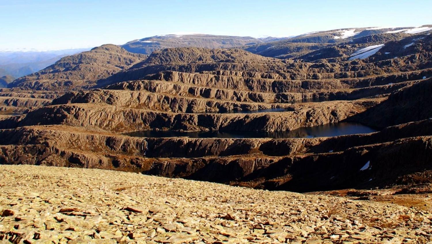Panoramautsikt over bratt, steinete fjellandskap med små innsjøar og snøflekker under klar blå himmel..