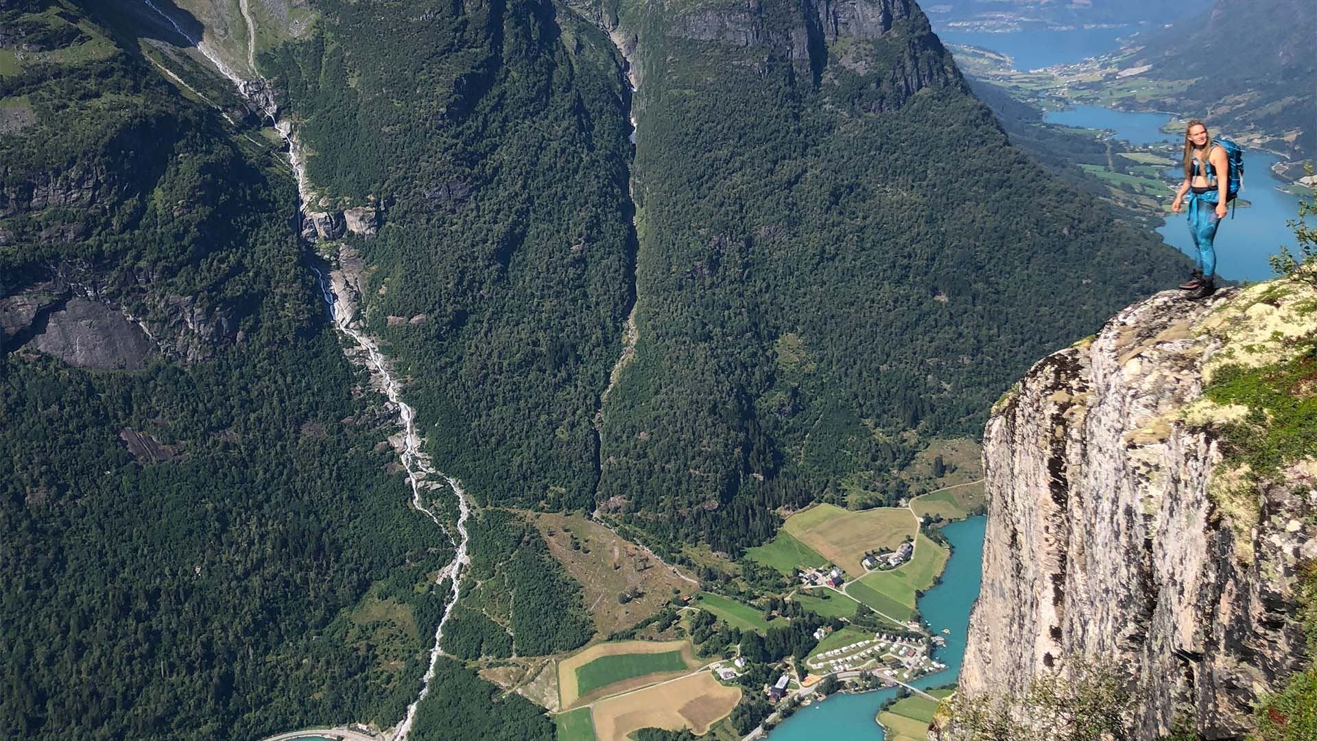 A hiker with a backpack stands on a rocky cliff edge overlooking a forested valley, river, farms, and blue fjords..
