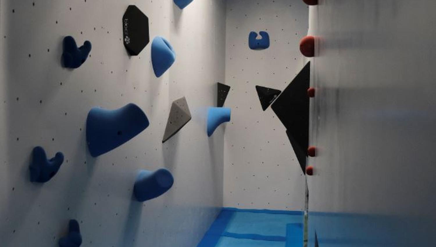 Narrow indoor bouldering corridor with blue climbing holds and volumes on white walls, blue floor mats, and a ceiling light..