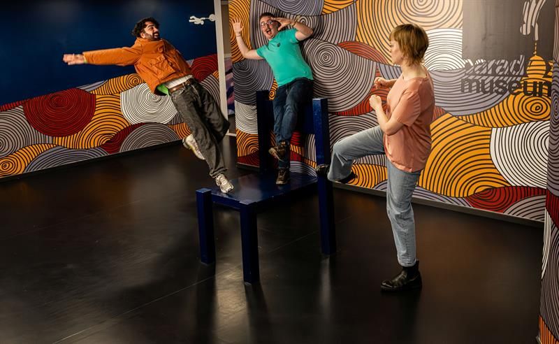Three people pose playfully in an interactive museum installation that creates the illusion of losing balance.