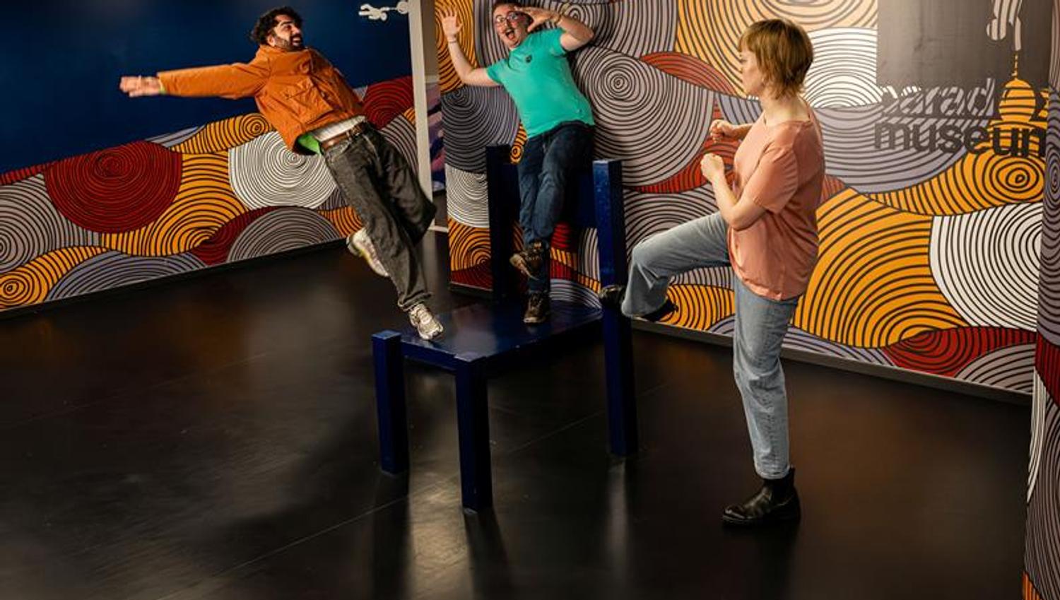 Three people pose playfully in an interactive museum installation that creates the illusion of losing balance.