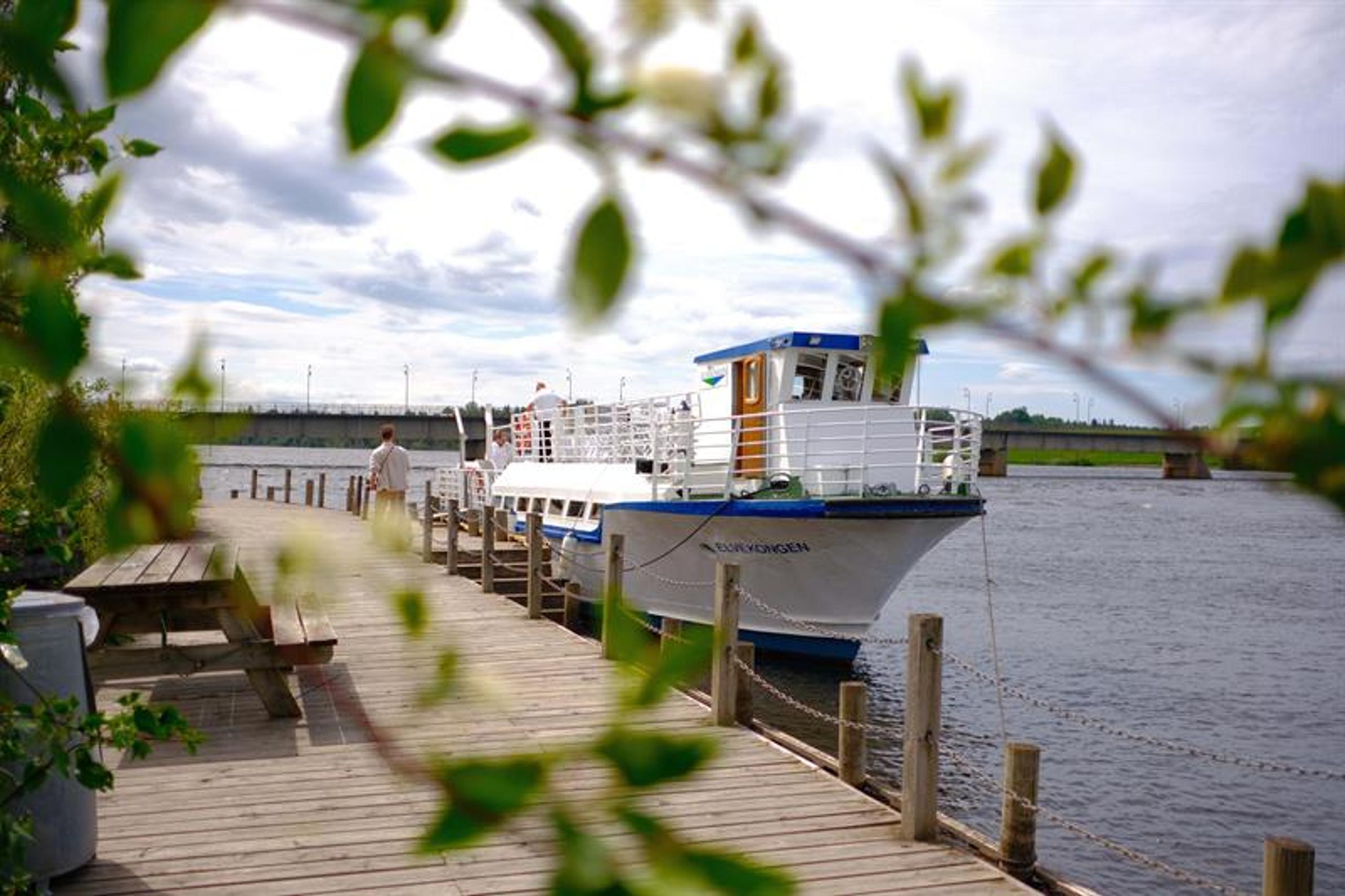 Brygge ved vannet med benker og planter i forgrunnen, og en båt i bakgrunnen