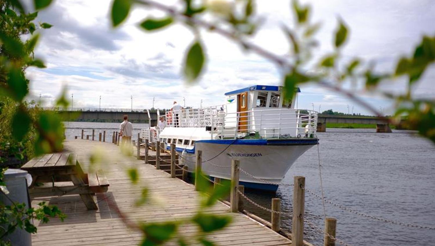 Brygge ved vannet med benker og planter i forgrunnen, og en båt i bakgrunnen