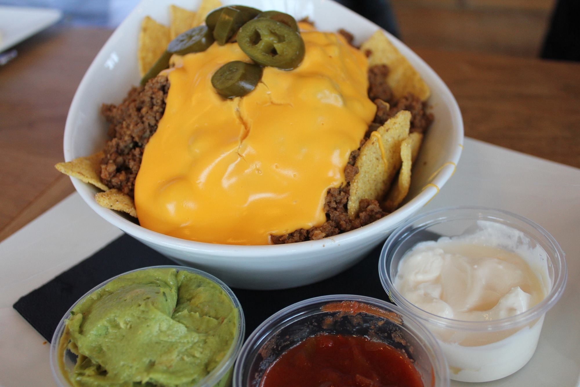 Hvit skål med nachos, kjøttdeig og smeltet ost toppet med jalapeño, med guacamole, salsa og rømme ved siden av..