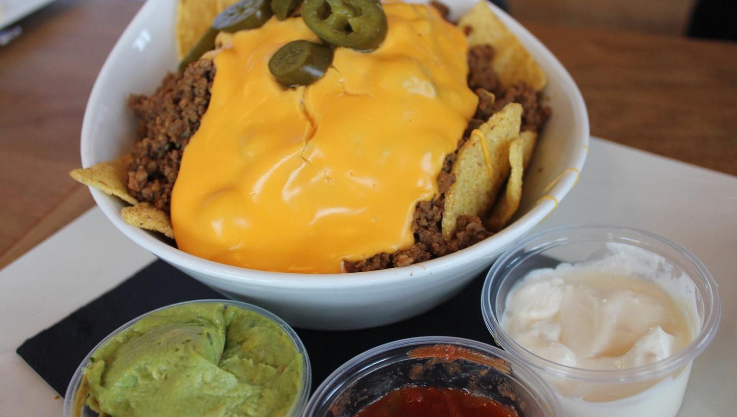 Hvit skål med nachos, kjøttdeig og smeltet ost toppet med jalapeño, med guacamole, salsa og rømme ved siden av..