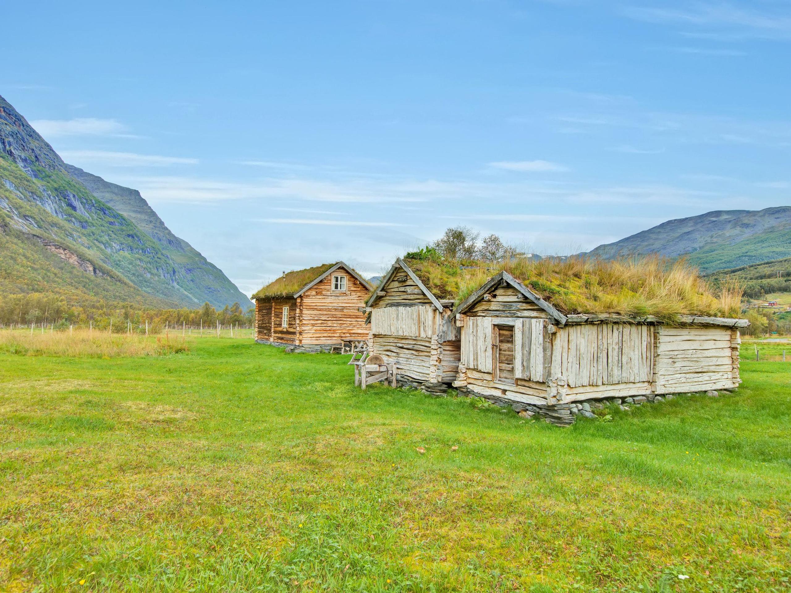 Three small, old wooden houses with grass on the roof. In the foreground cut grass and in the background blue sky and mountains