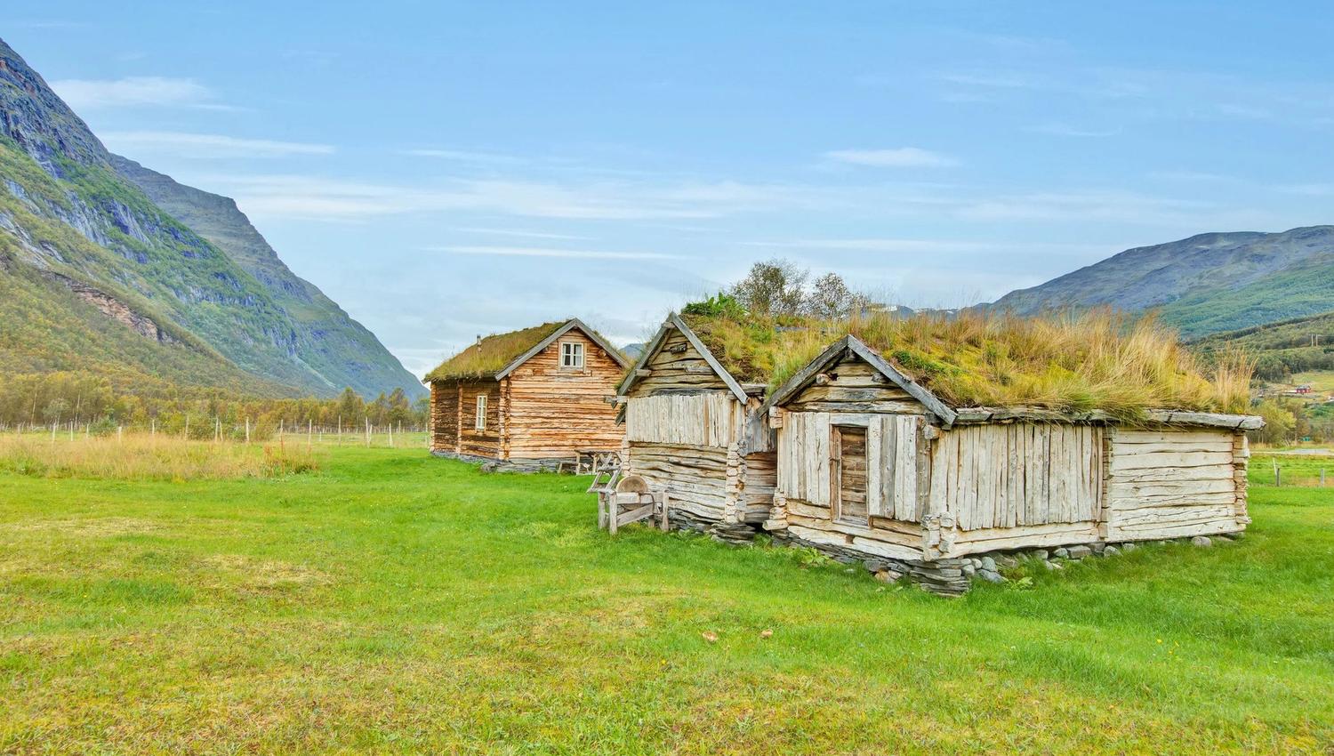 Three small, old wooden houses with grass on the roof. In the foreground cut grass and in the background blue sky and mountains