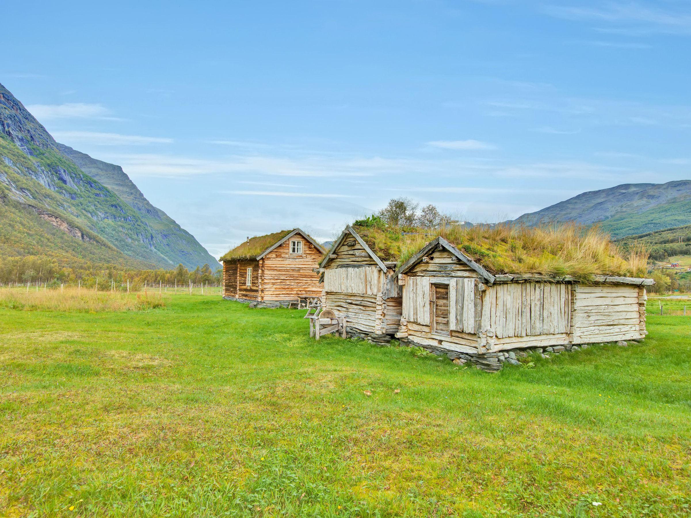 Three small, old wooden houses with grass on the roof. In the foreground cut grass and in the background blue sky and mountains