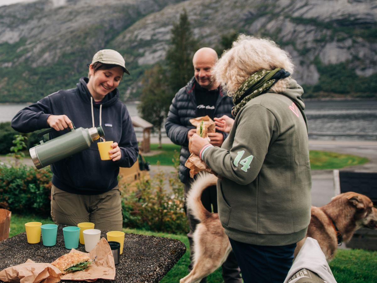 Tre personer deler mat og drikke utendørs ved en fjord, med fjell i bakgrunnen og hunder i nærheten..