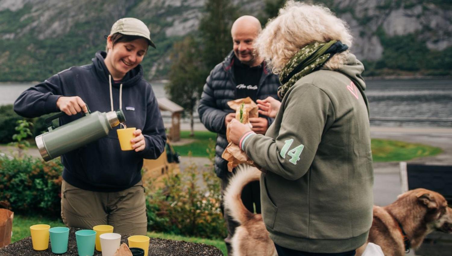 Tre personer deler mat og drikke utendørs ved en fjord, med fjell i bakgrunnen og hunder i nærheten..