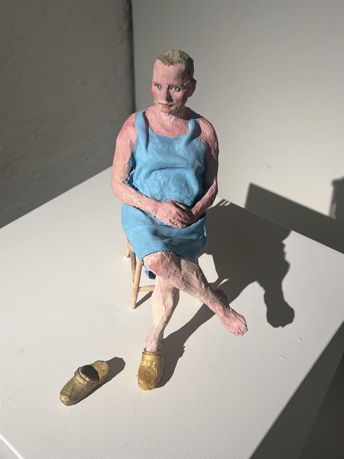 Sculpture of a seated figure in a blue dress with shoes placed on the floor in front.