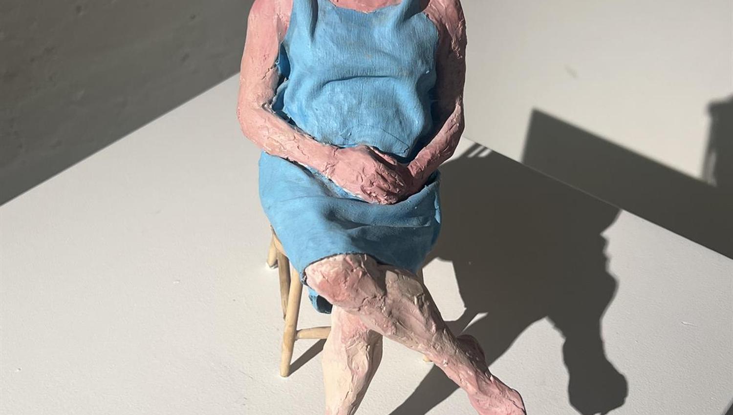 Sculpture of a seated figure in a blue dress with shoes placed on the floor in front.