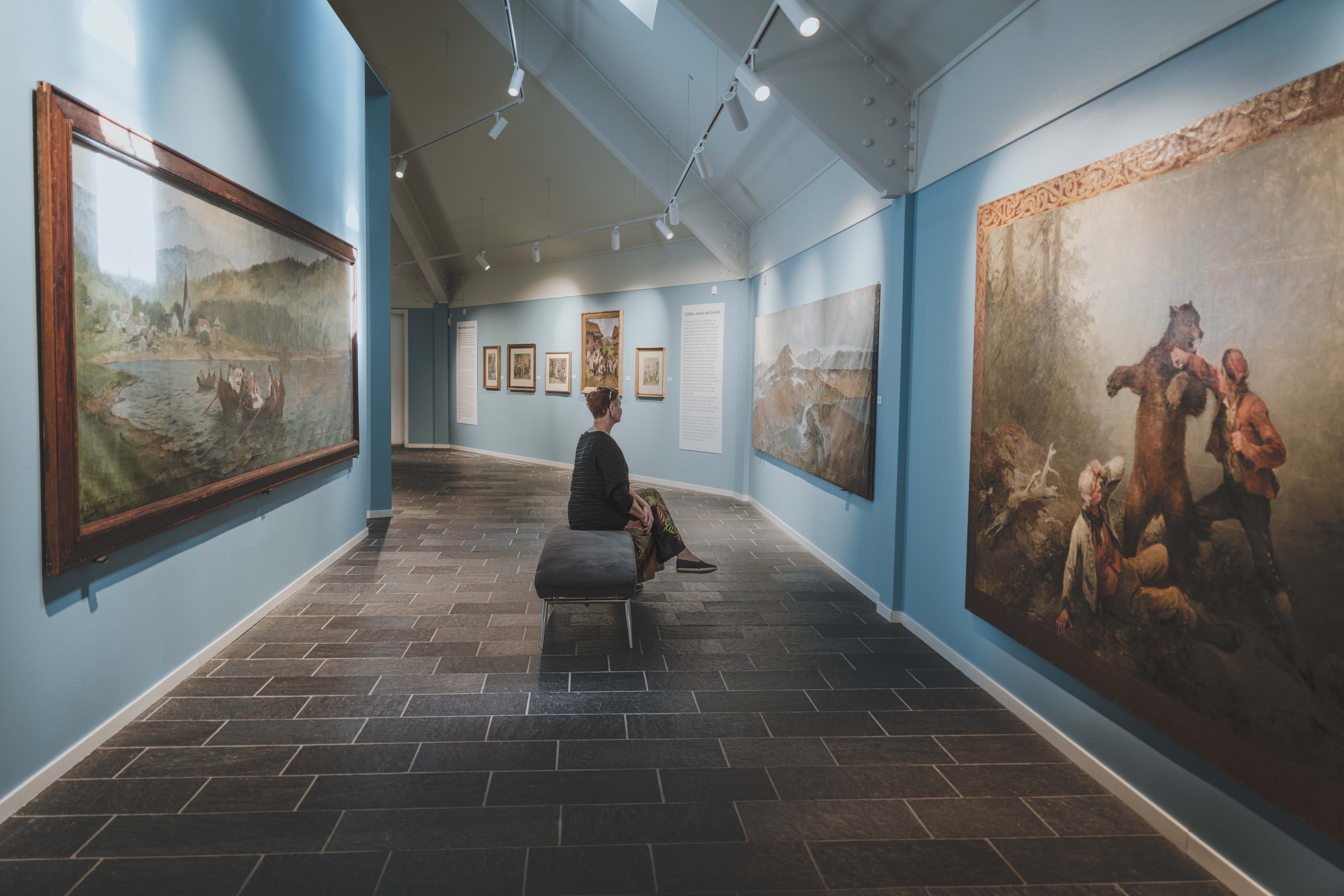 A woman is sitting on a bench viewing the paintings.