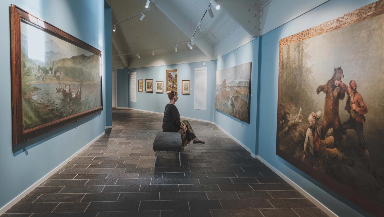 A woman is sitting on a bench viewing the paintings.