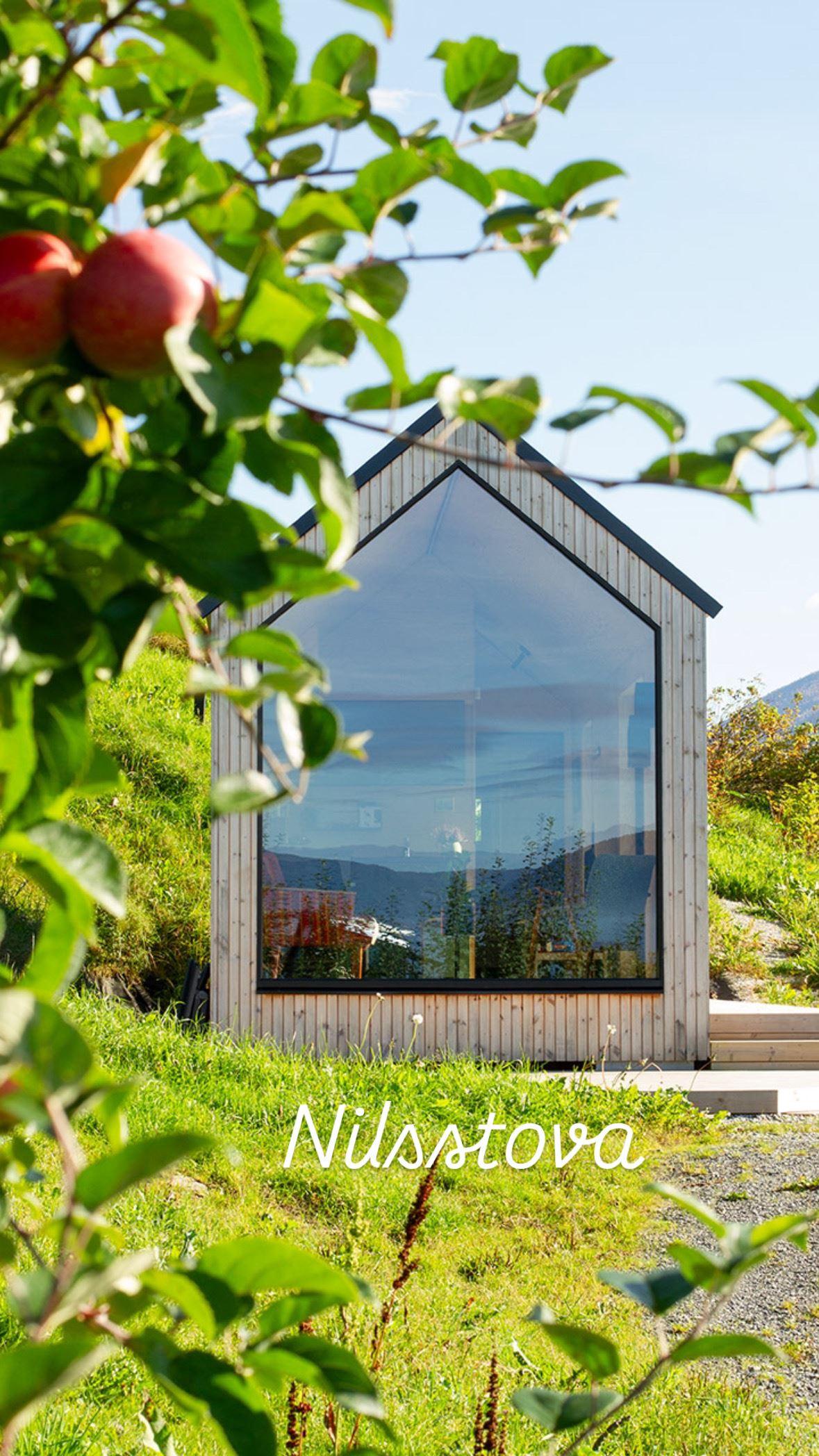 Small wooden cabin with a tall glass front sits on a grassy hillside, framed by leafy branches with apples..