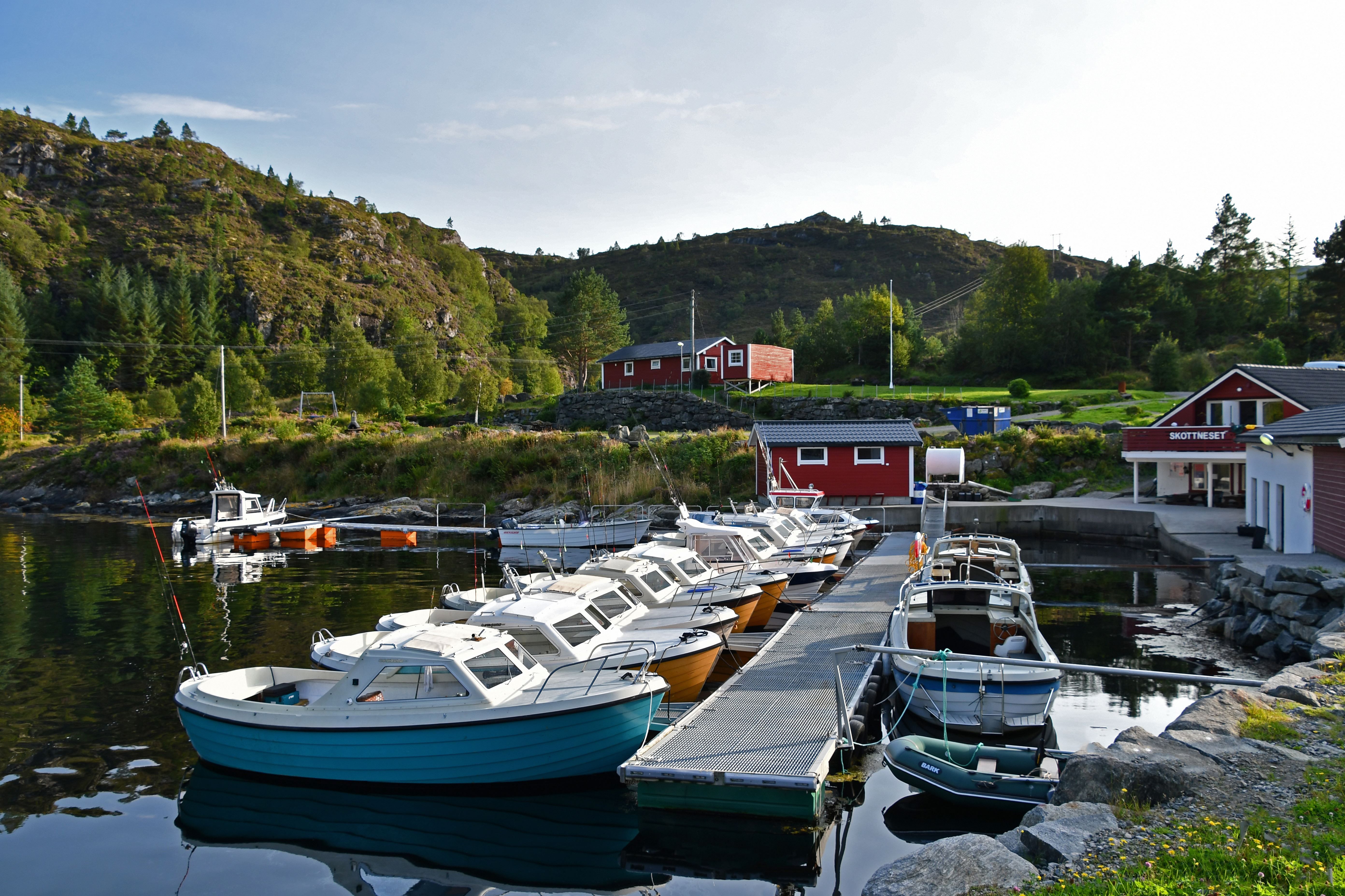 Several small motorboats tied to a floating dock in a calm inlet, with red buildings and green hills behind..