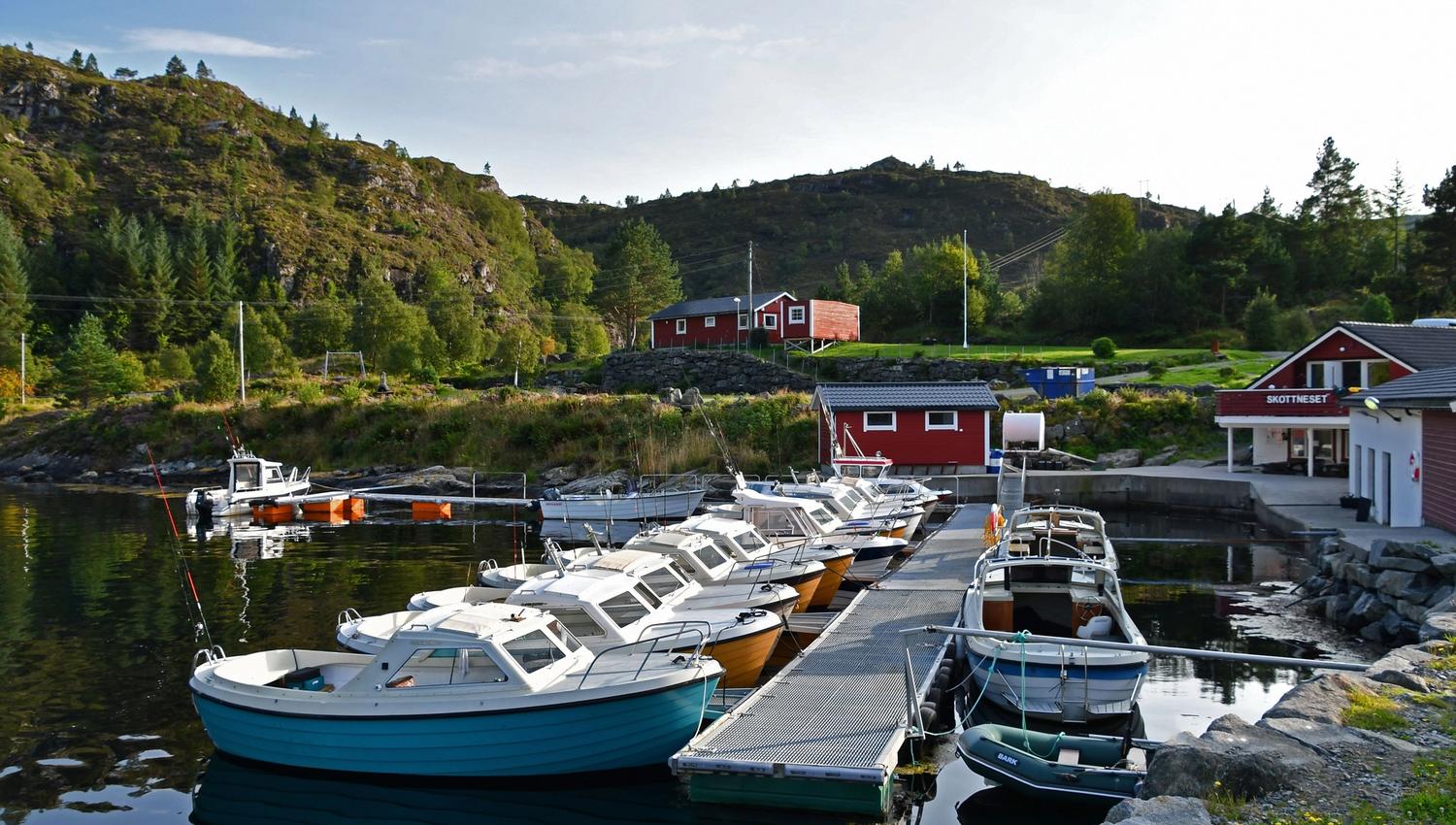Several small motorboats tied to a floating dock in a calm inlet, with red buildings and green hills behind..