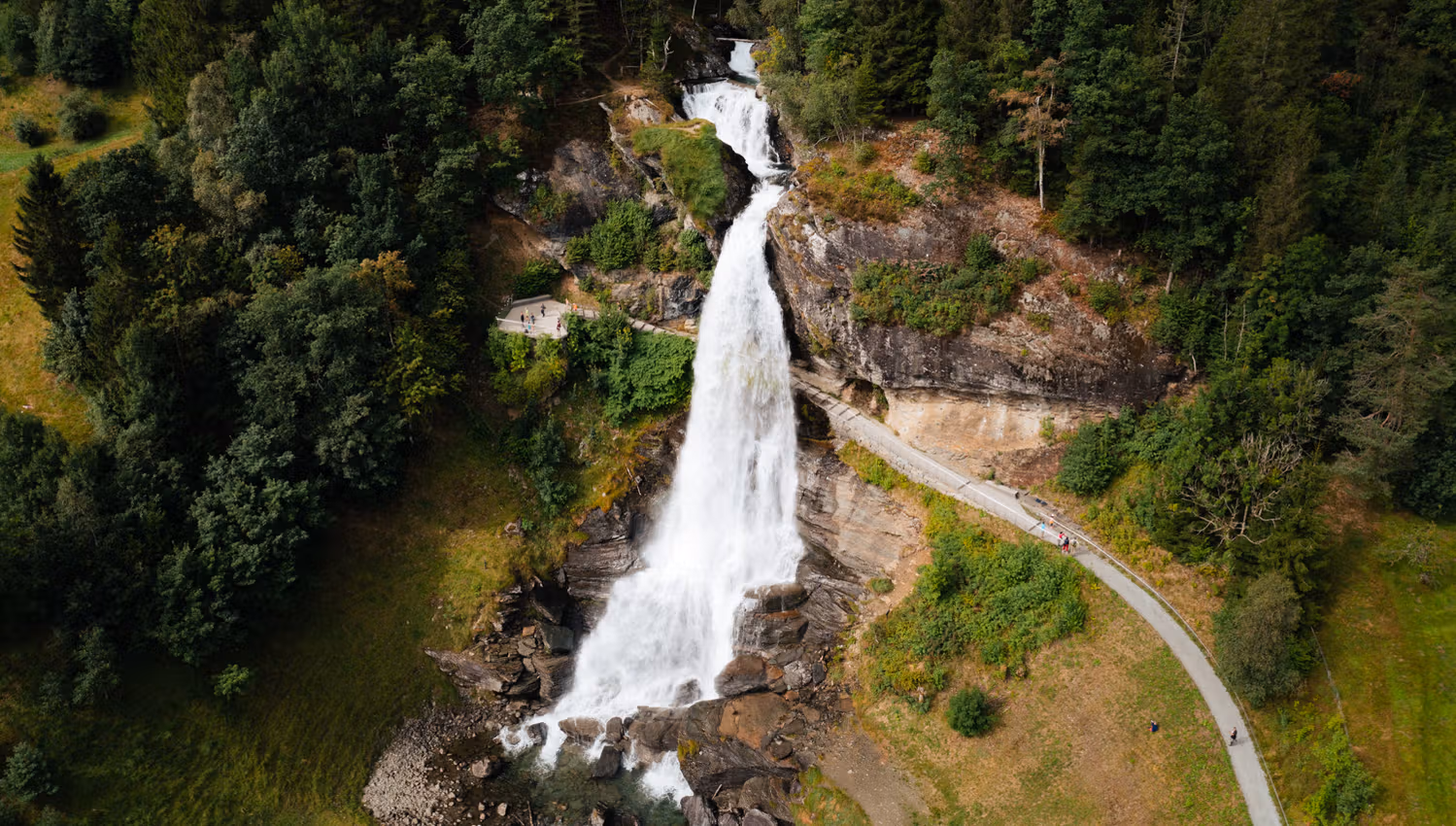 Aerial view of Steinsdalsfossen, a majestic waterfall surrounded by lush greenery and a walking path, with people strolling close to the falls in Norw