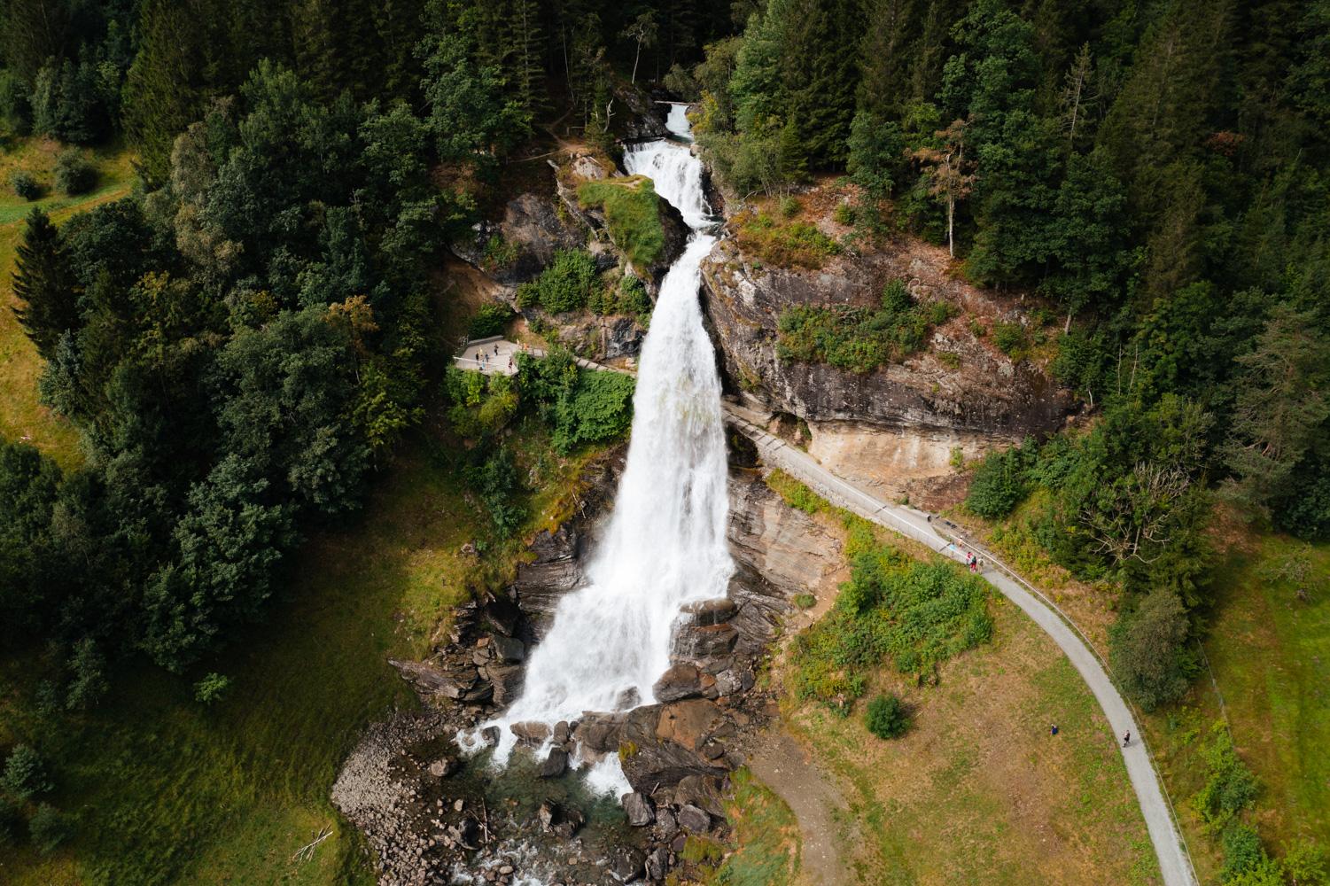 Aerial view of Steinsdalsfossen, a majestic waterfall surrounded by lush greenery and a walking path, with people strolling close to the falls in Norw