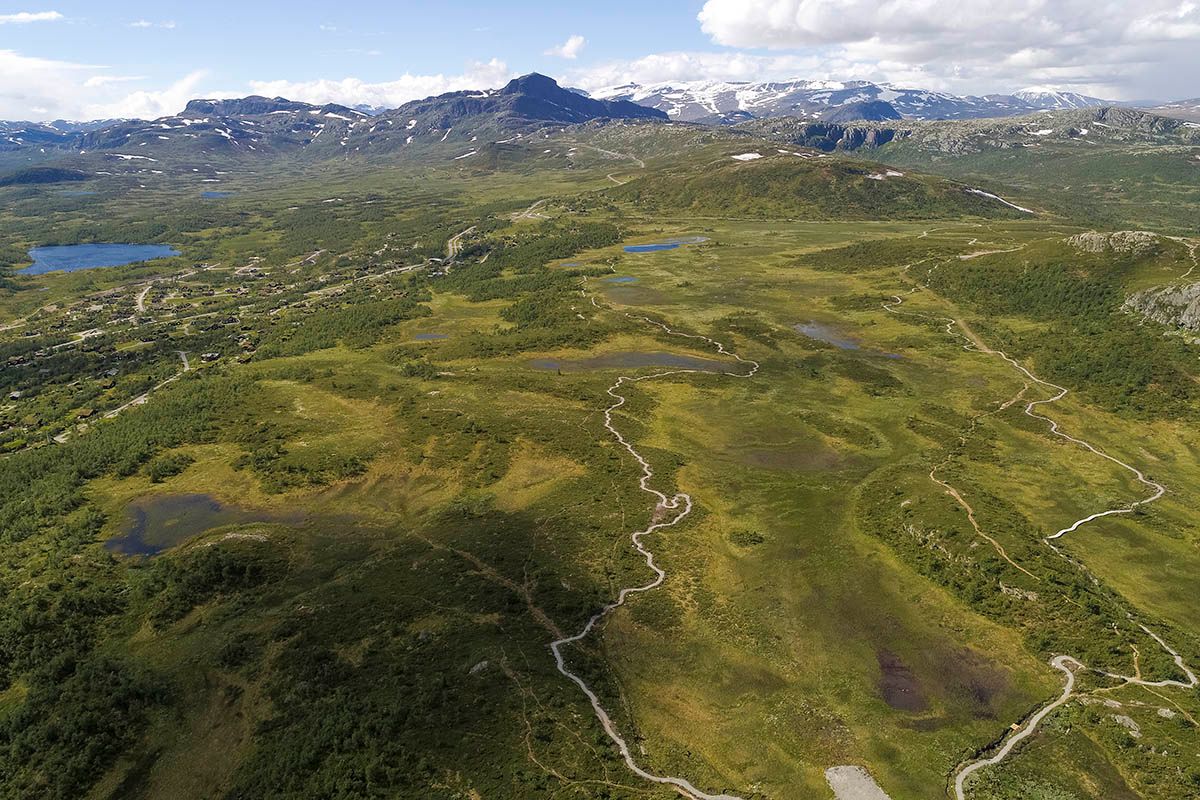 Aerial image of a mountain plateau with a gravel trail for terrain cyclists leading around a swamp. High mountains in the background.