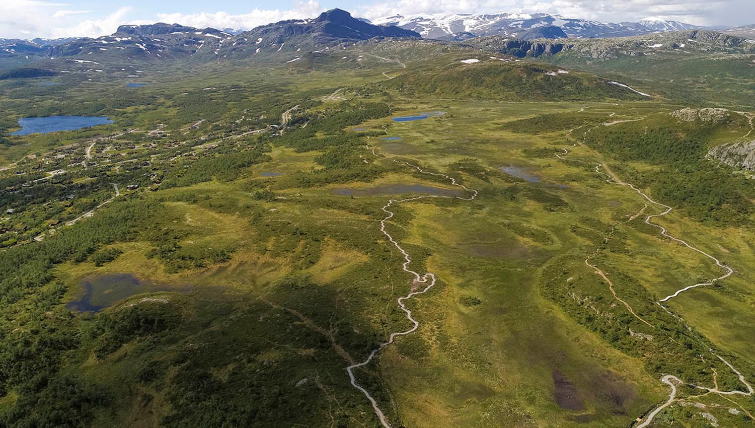 Aerial image of a mountain plateau with a gravel trail for terrain cyclists leading around a swamp. High mountains in the background.