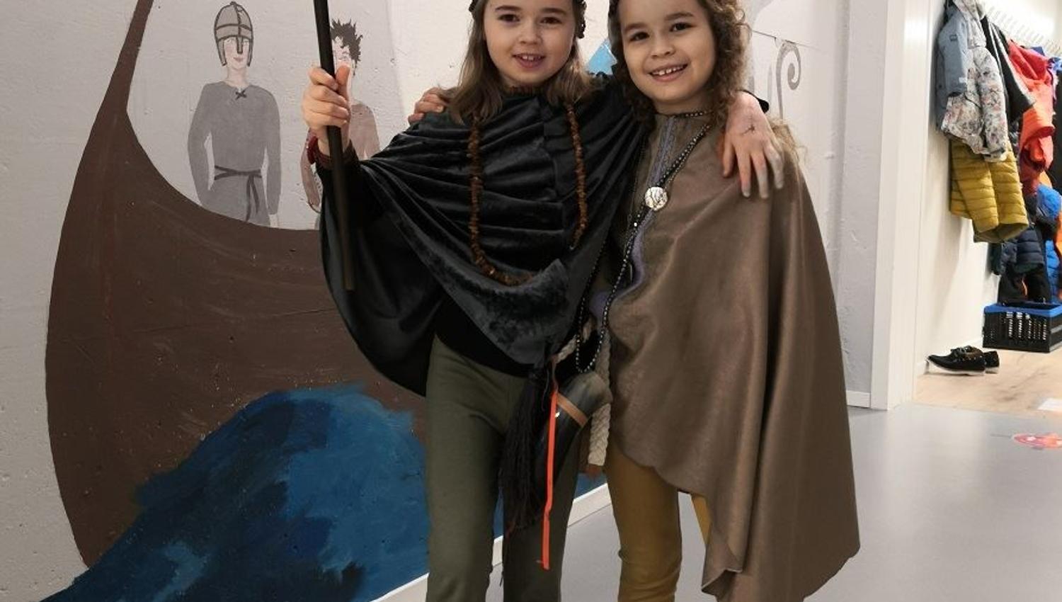 Two people in cloak costumes pose with arms around each other beside a Viking ship wall mural in a hallway..
