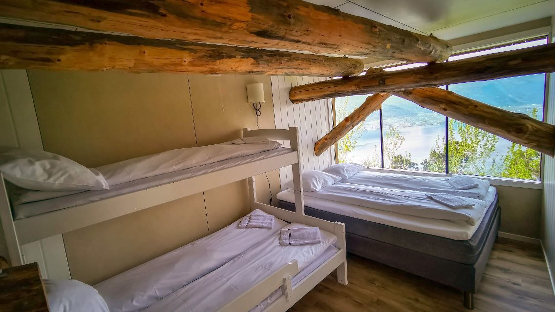 Rustic bedroom with bunk beds and a double bed, exposed log beams overhead, and large windows overlooking a lake..