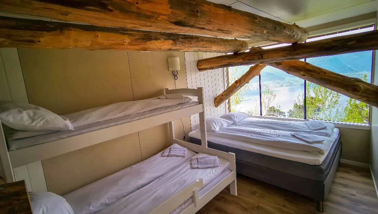 Rustic bedroom with bunk beds and a double bed, exposed log beams overhead, and large windows overlooking a lake..