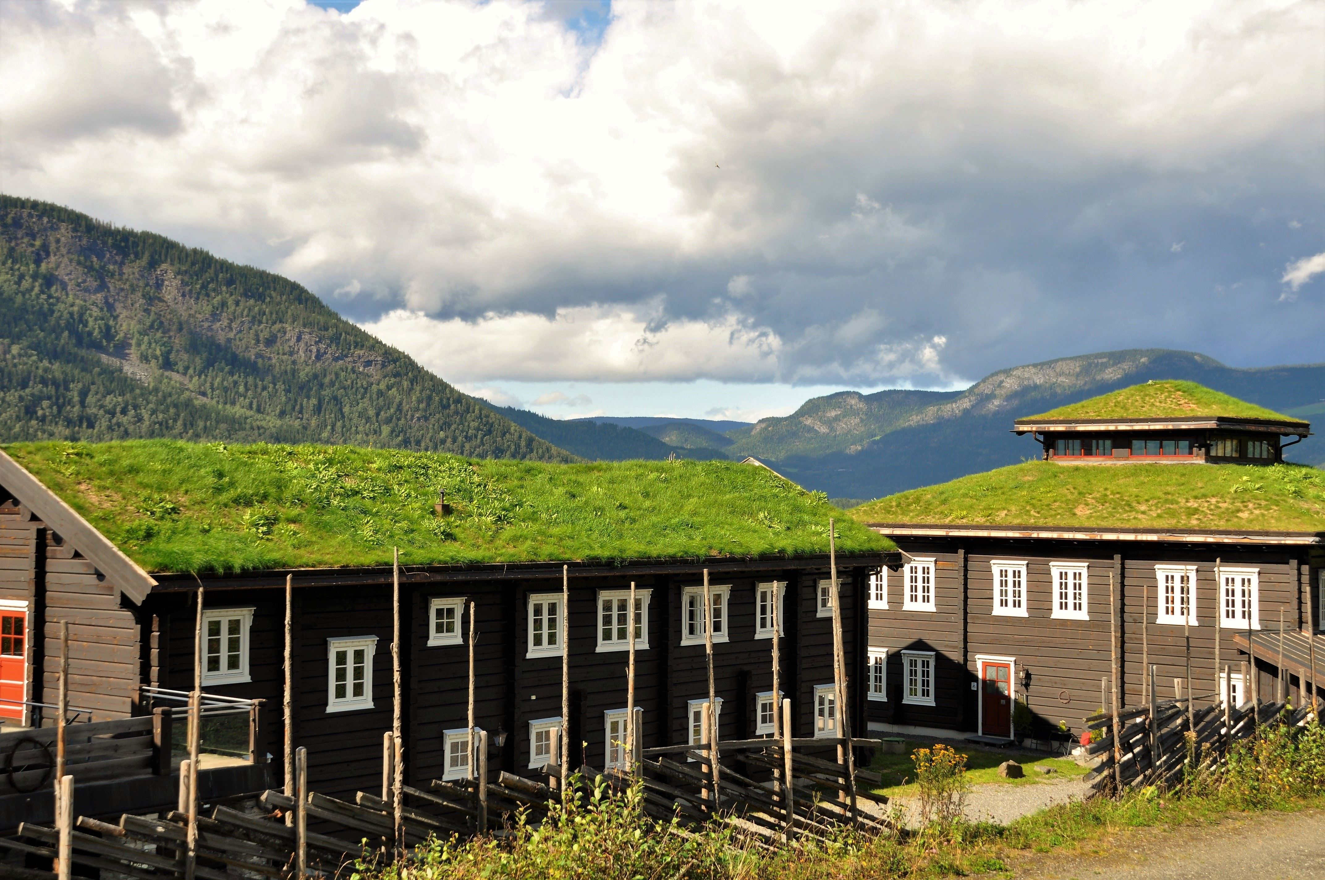 Mörka träbyggnader med grästak i fjällmiljö, med berg i bakgrunden och stora moln på himlen.