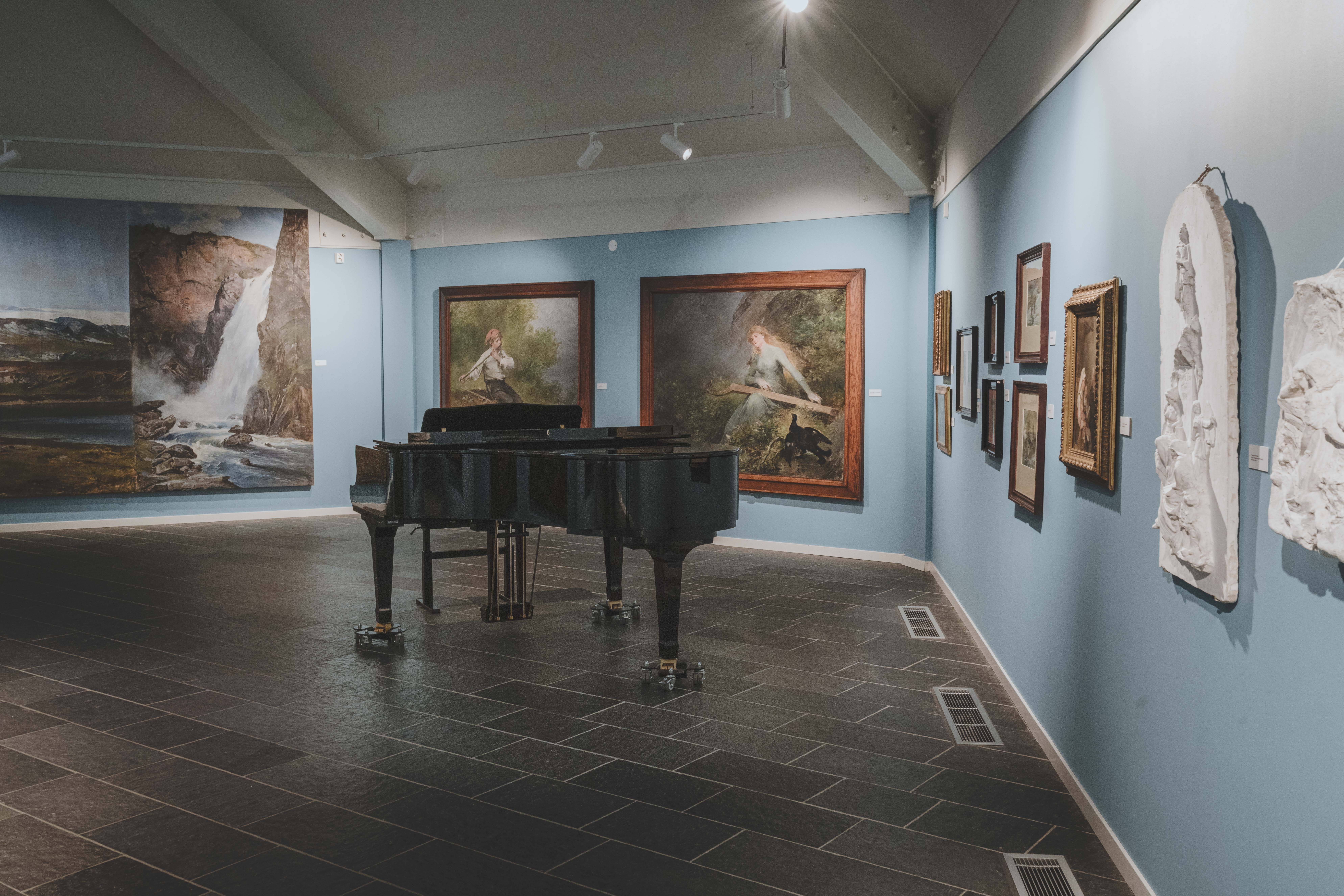 A baby grand piano in front of some of Bergsliens paintings.