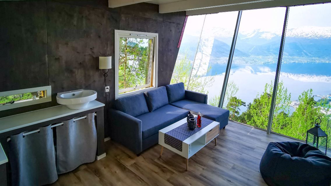Modern living room with sofa, coffee table, sink counter, and large slanted windows overlooking a lake and mountains..