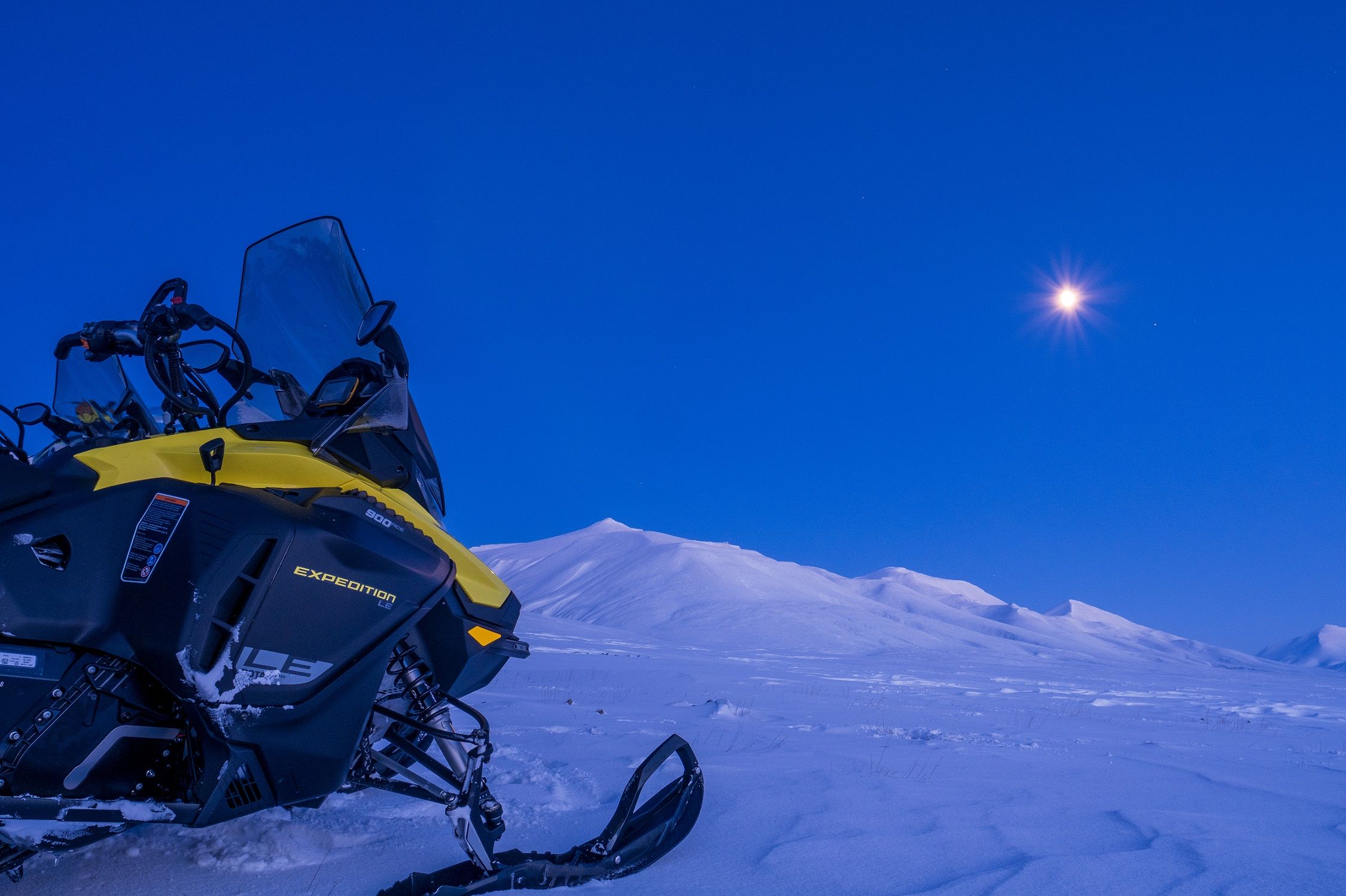 A snowmobile in a snow-covered landscape covered in blue light with a bright moon in the sky